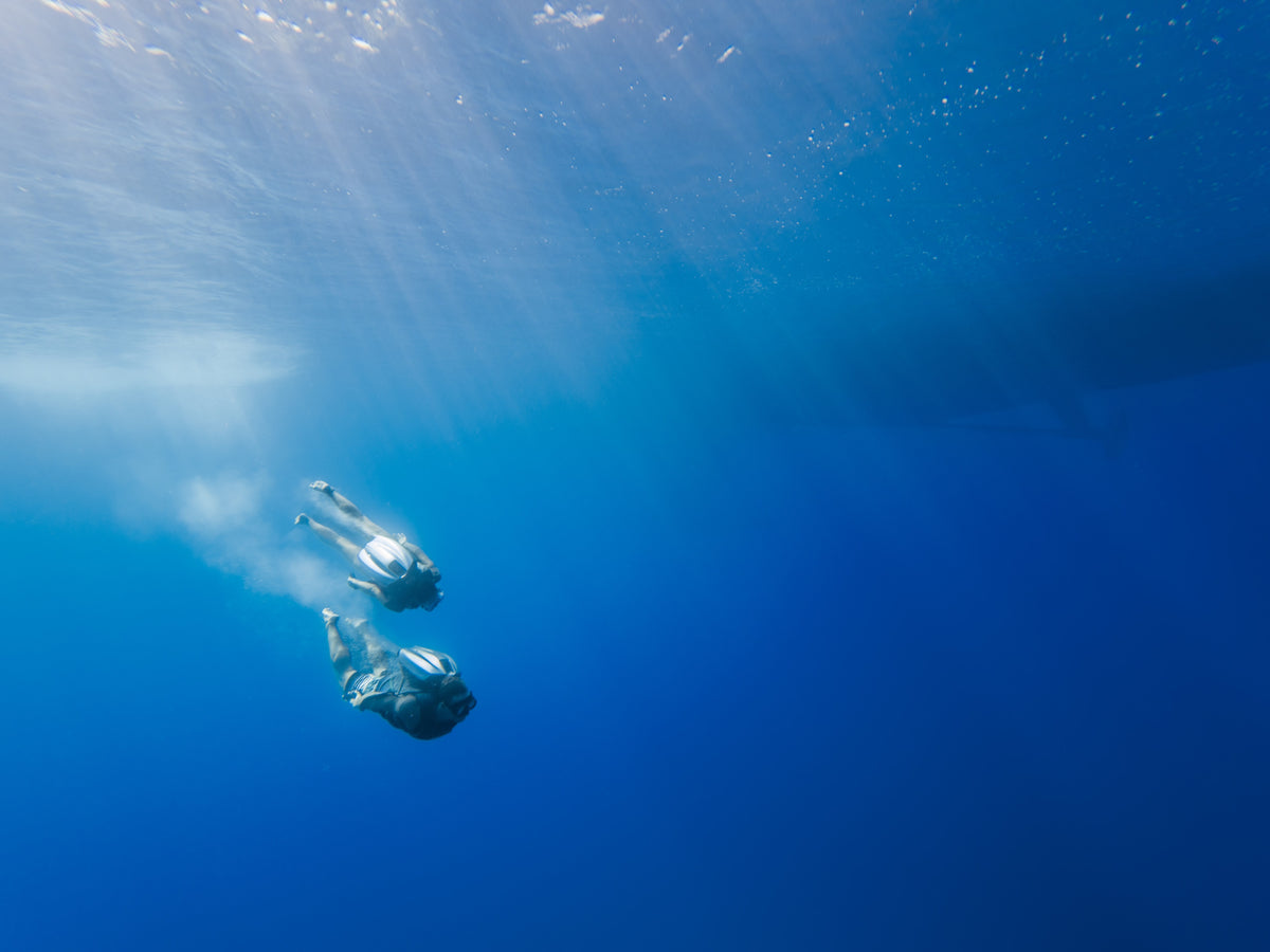 Underwater ocean scene with two CudaJet users in clear blue water, with sunlight filtering down from the surface and a large boat visible in the background.