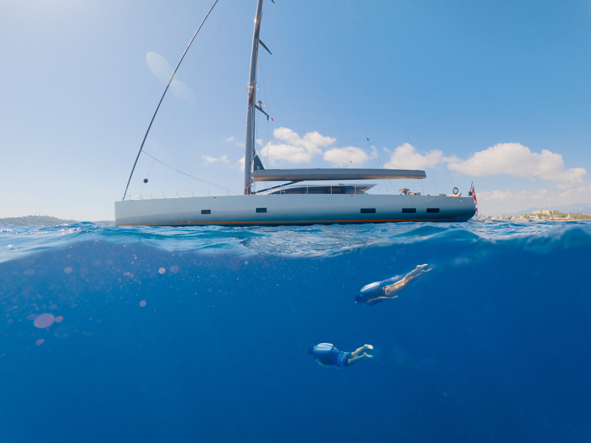 A large silver sailboat floating on the surface of deep blue ocean water under a bright blue sky, with two people Jetpacking below the waterline.