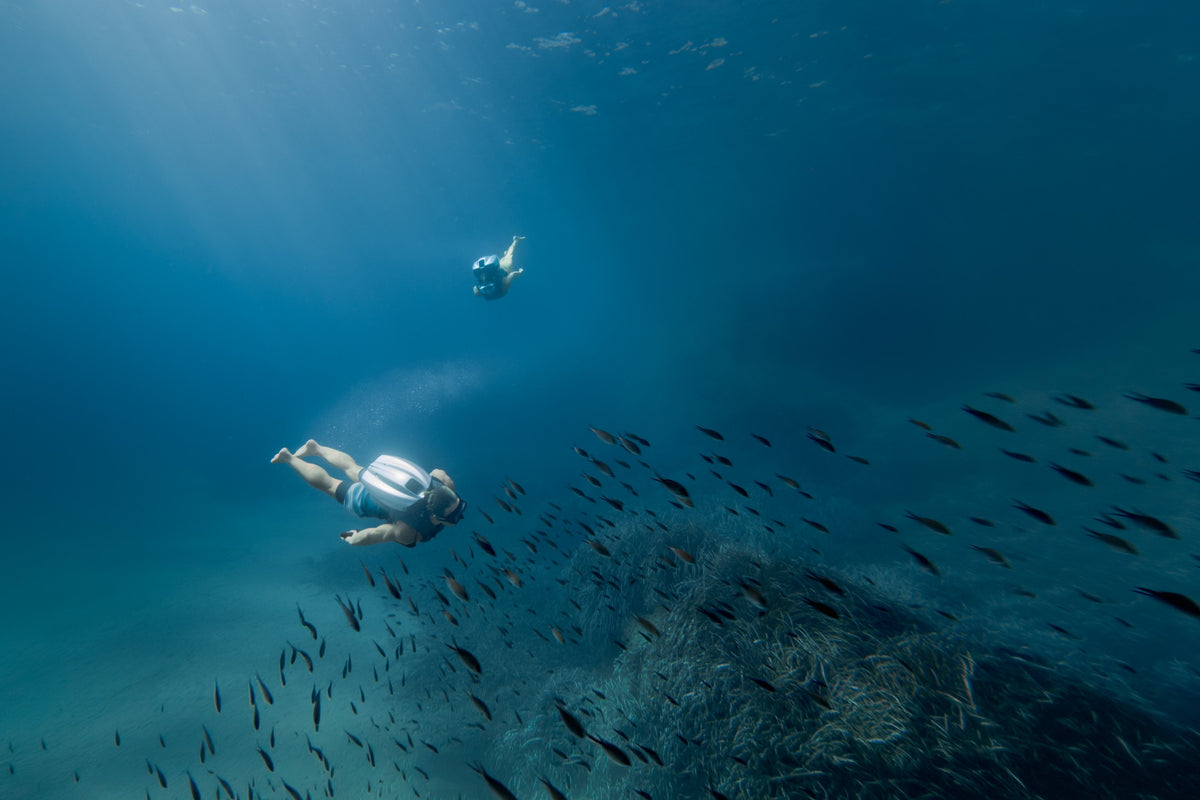 An underwater scene with divers wearing underwater jetpacks near a coral reef while surrounded by a school of small fish, with another diver visible in the distance under bright sunbeams filtering through the water.