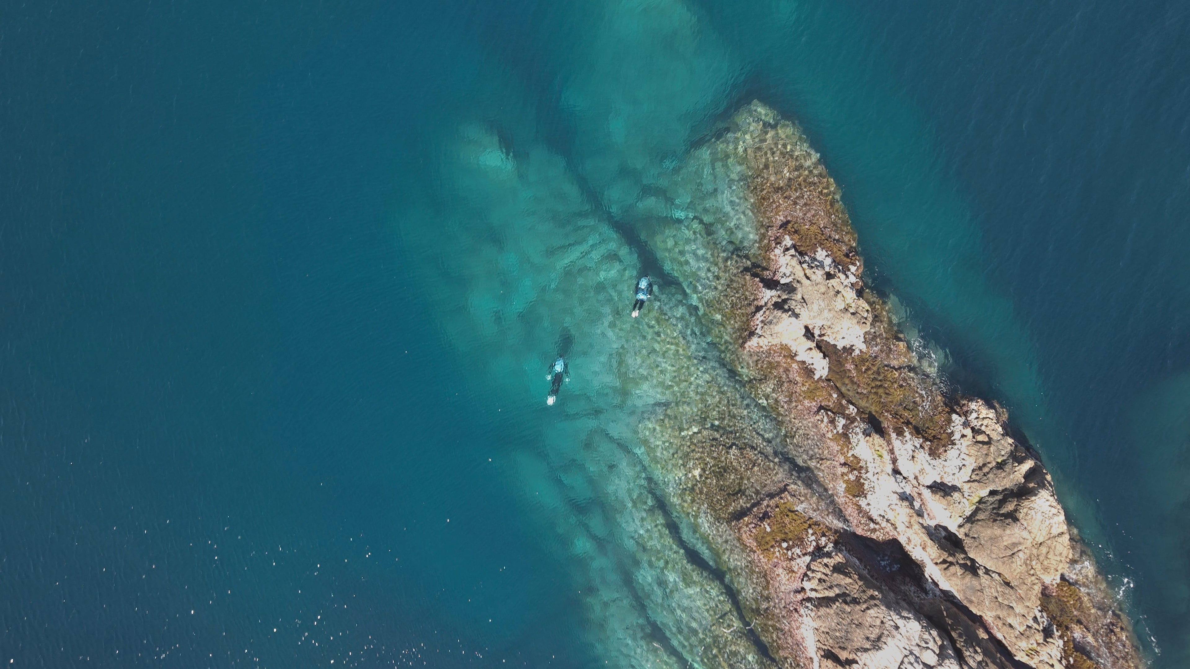 Aerial view of a  Jetpacks flying round a rocky coastline with clear blue water.