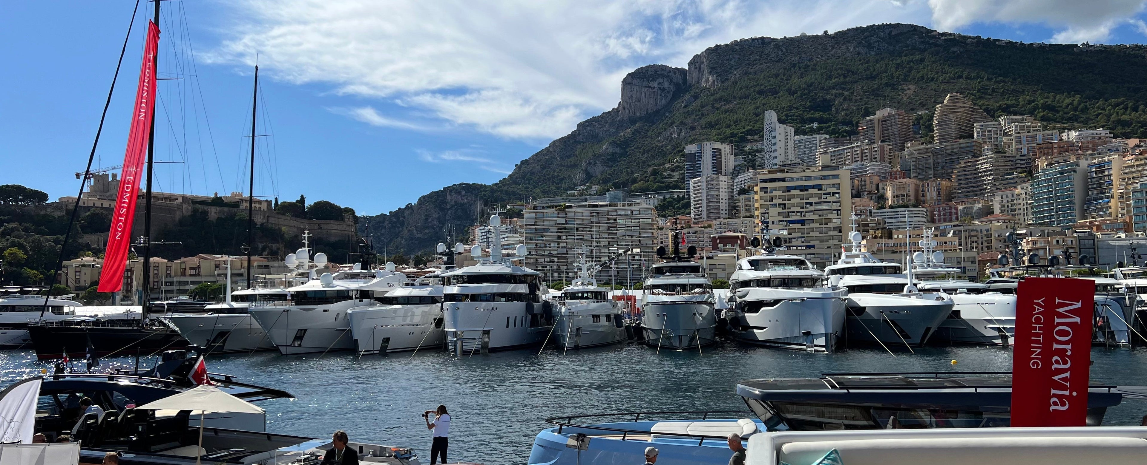 A scenic view of a marina filled with white yachts and boats, set against a backdrop of a steep green mountain and dense white buildings under a blue sky with clouds. In the foreground, a white yacht deck with wooden railings and a person standing is visible.