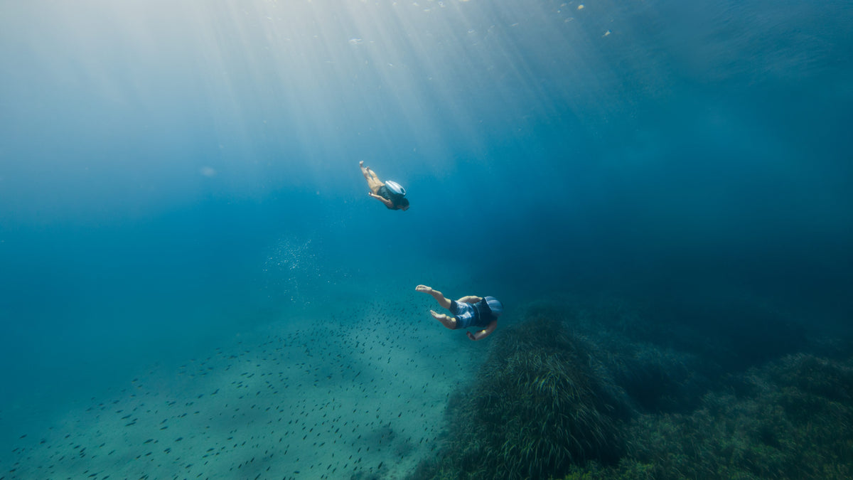 Underwater scene with two divers wearing CudaJet Jetpacks in clear blue water, with sun rays filtering down from the surface and a seabed with marine growth below.