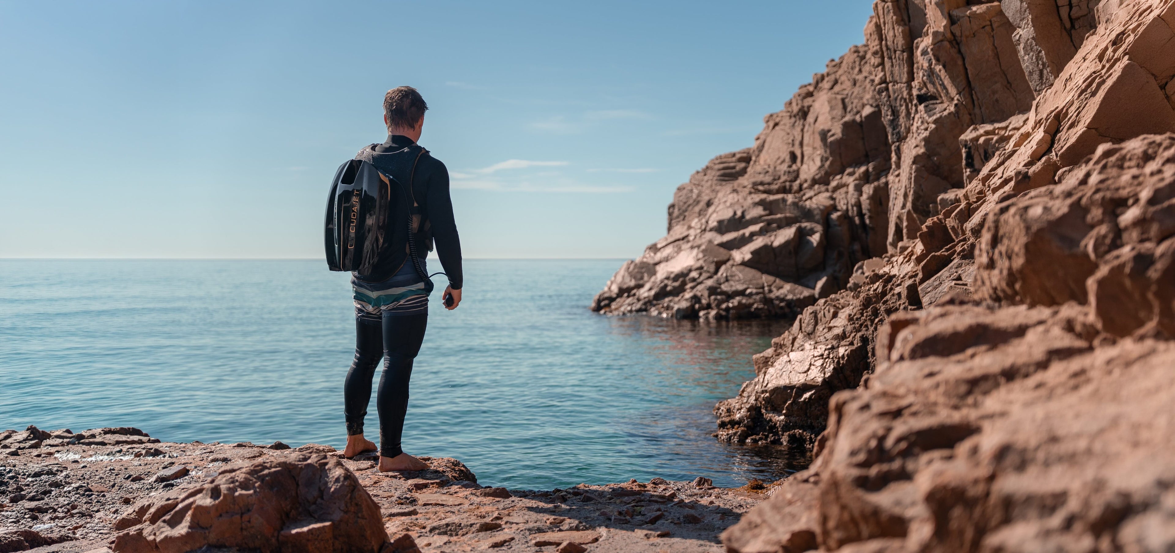 Person wearing a Cudajet underwater jetpack on a rocky coastline looking out at the ocean.